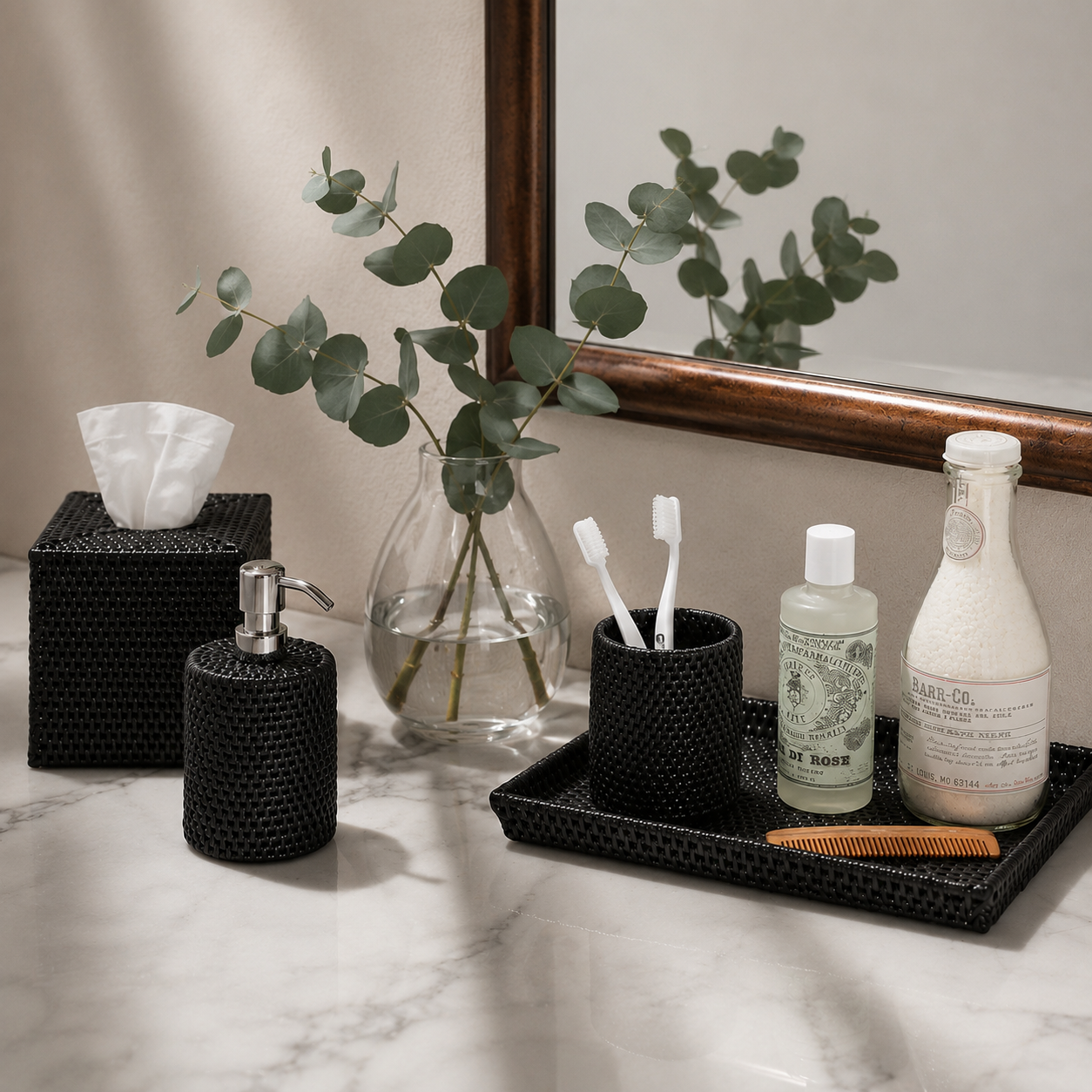 A styled bathroom vanity scene with black woven-texture accessories including a tissue box cover, soap dispenser, and cup on a tray, alongside a glass bottle and a comb. A mirror, marble countertop, and eucalyptus in a vase are visible in the background.
