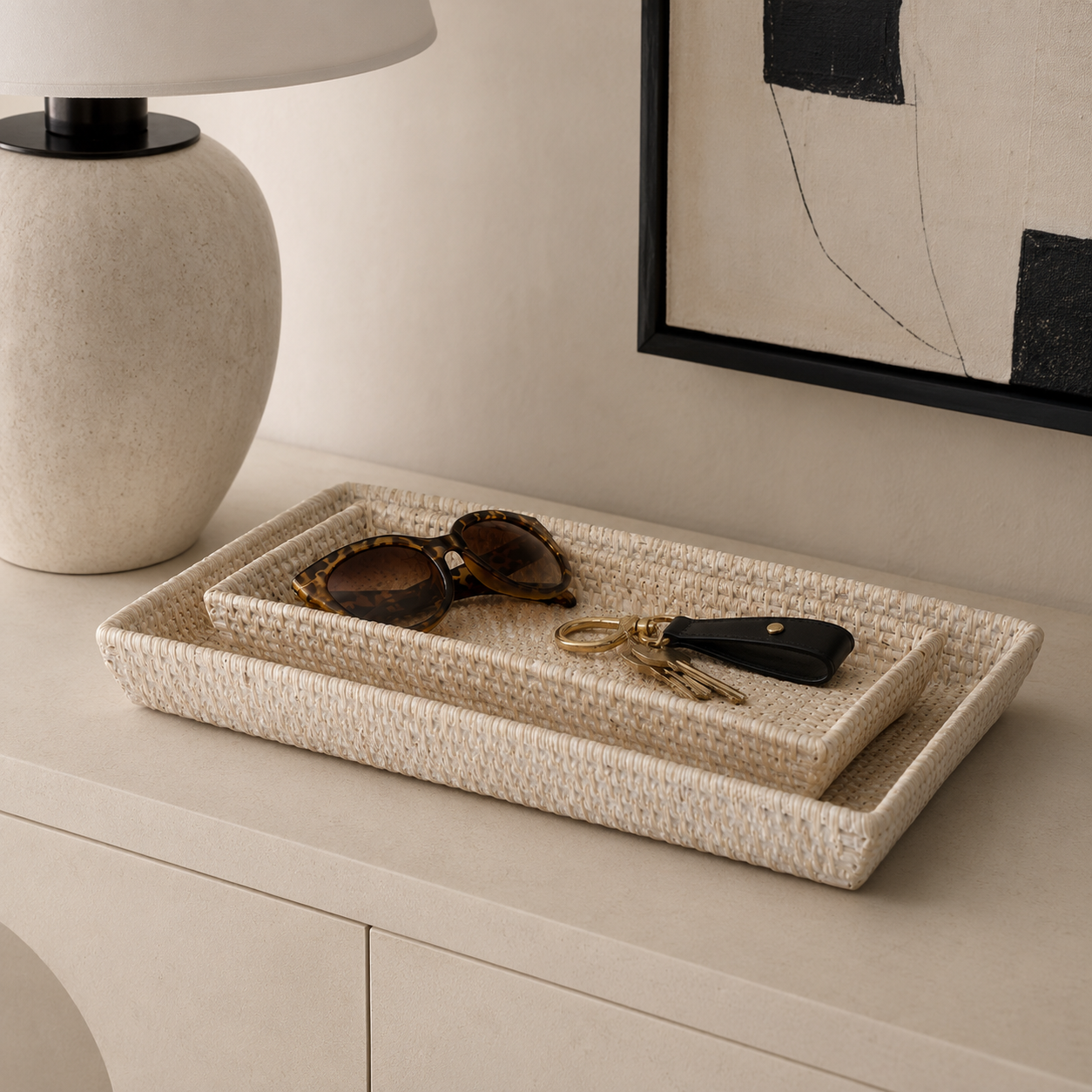 Two nested woven rectangular trays in a light natural fiber material, styled on a cream-colored console table with a lamp, sunglasses, and keys. A framed abstract print hangs on the wall in the background.