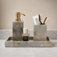 A marble bathroom accessory set on a tray, including a soap dispenser with a gold pump, a toothbrush holder with two toothbrushes, a small Acqua di Parma bottle, and a white tube, arranged on a marble surface against a beige wall.