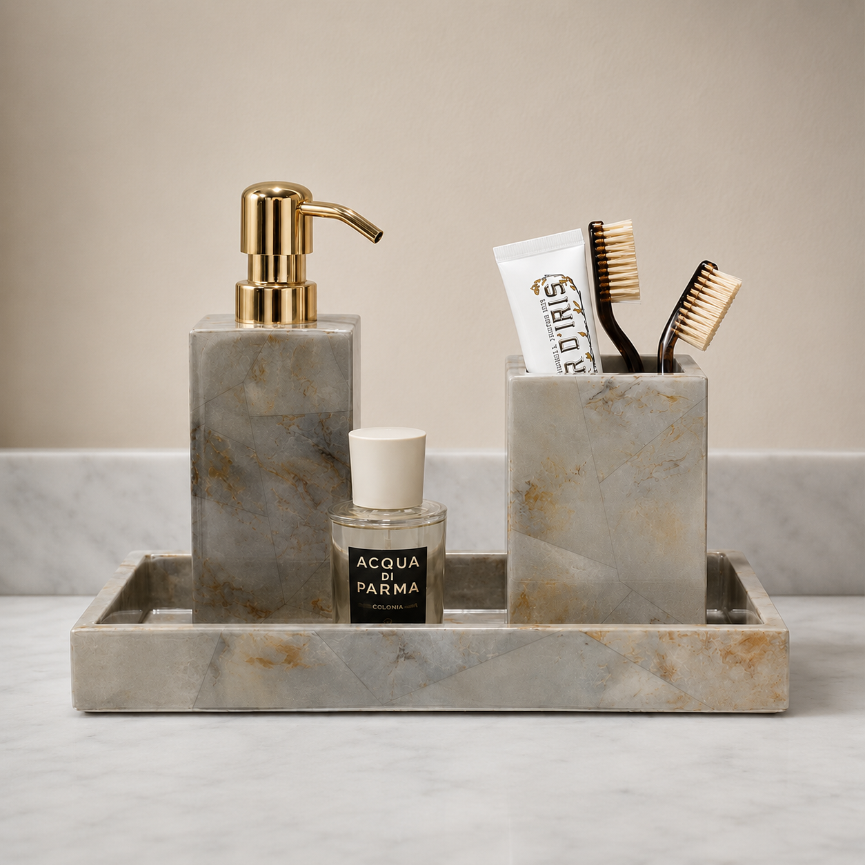 A marble bathroom accessory set on a tray, including a soap dispenser with a gold pump, a toothbrush holder with two toothbrushes, a small Acqua di Parma bottle, and a white tube, arranged on a marble surface against a beige wall.