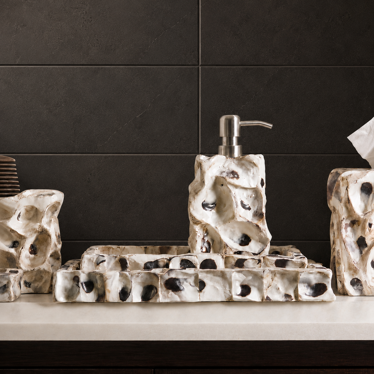 A bathroom vanity set featuring a soap dispenser, tray, and tissue holder in a textured, organic white ceramic style with black speckles, set against a dark charcoal tiled wall.