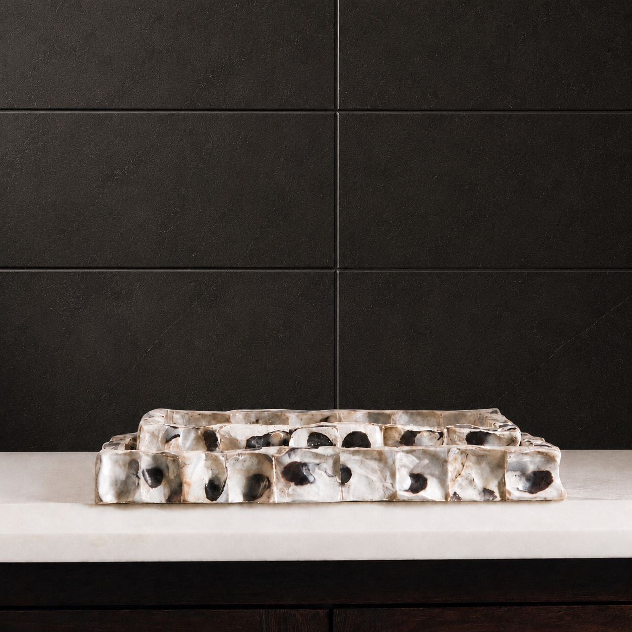 A rectangular ceramic tray with a stacked, tiered design, featuring a textured, organic pattern in white, beige, and black. The tray is displayed on a light-colored stone countertop against a dark charcoal tiled wall.