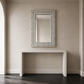 A rectangular wall mirror with an ornate silver mosaic frame, mounted on a beige wall above a white console table.