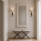 A tall rectangular wall mirror with a textured white frame hangs on a beige wall in a hallway, flanked by two black wall sconces above two matching white-and-black stools.
