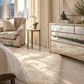 A styled bedroom corner featuring a fluffy cream shag rug, a mirrored dresser with gold trim, a patterned armchair, and a small side table near tall cream curtains and a window.