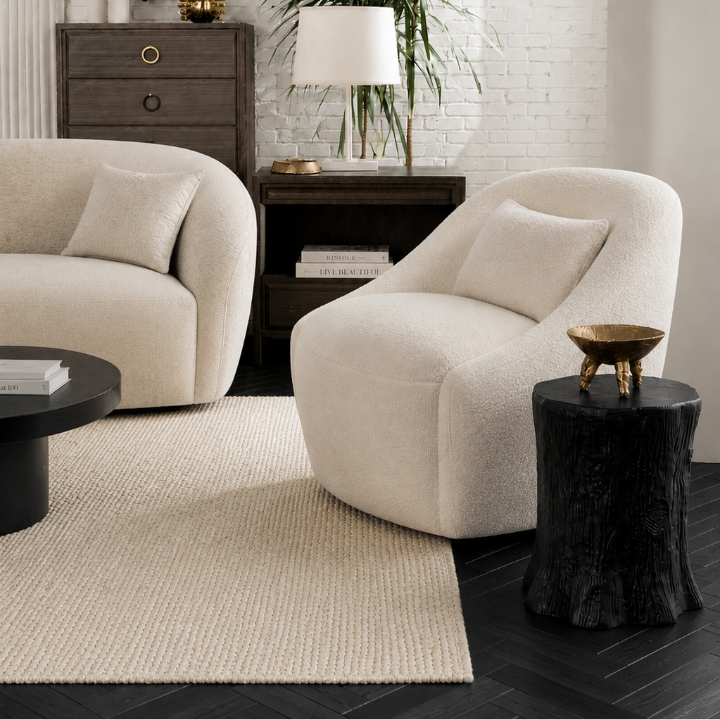 A styled living room featuring two cream boucle armchairs, a dark wood sideboard with a gold-framed mirror, a potted palm, and a textured cream rug on dark wood floors.