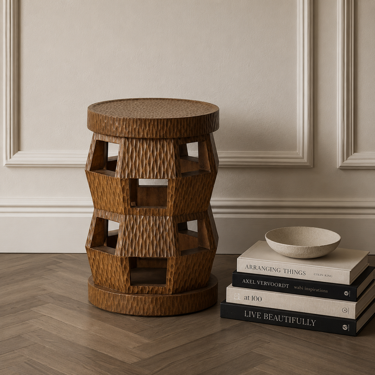 A round woven rattan accent stool in a warm medium brown tone, styled in a room setting next to a stack of books and a small ceramic bowl on a herringbone wood floor against a beige paneled wall.