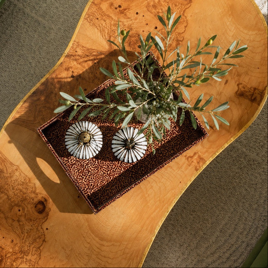 An organic-shaped wooden serving board or tray with dramatic burl wood grain, styled with a small potted olive branch and two white decorative pumpkins on a textured gray rug.