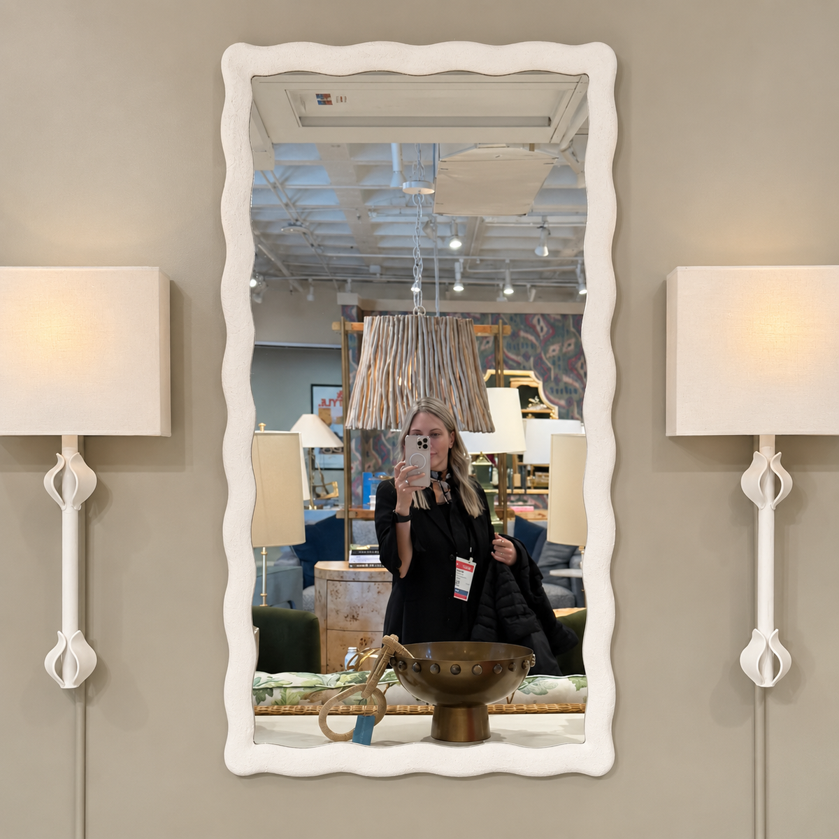 A vertical rectangular wall mirror with a wavy white frame, mounted on a beige wall between two matching white sconces. The mirror reflects a person taking a photo in a furniture store.