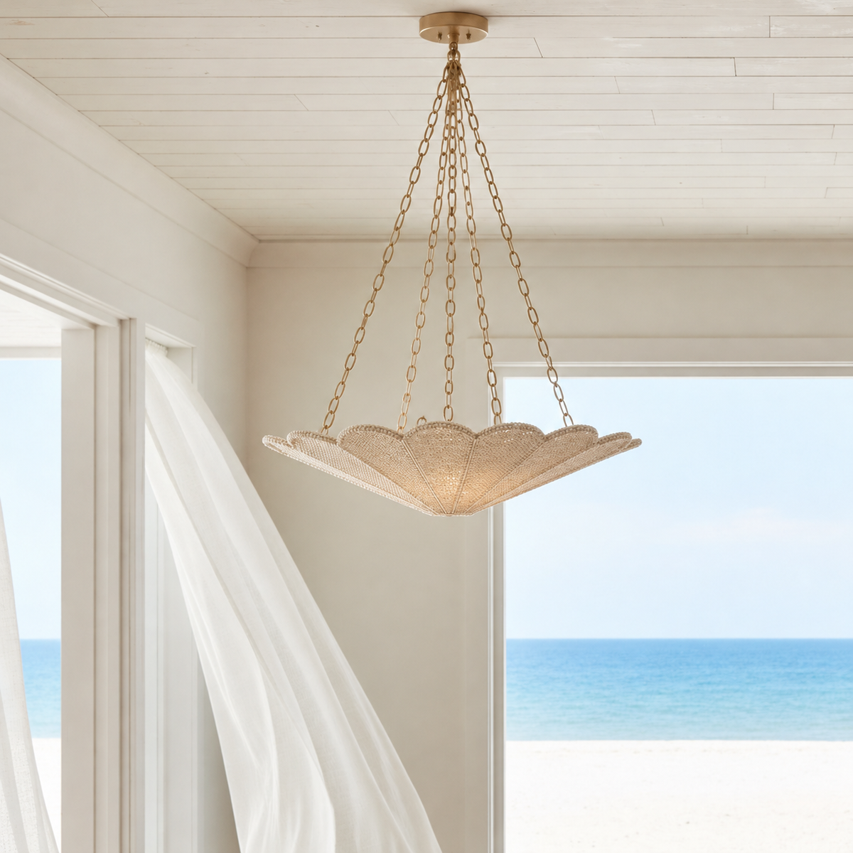 Sofia Chandelier featuring a sculptural scalloped ivory shade suspended by long antique brass chains from a round ceiling mount, styled in an airy coastal interior with white shiplap ceilings, flowing sheer curtains, natural daylight, and serene ocean views.
