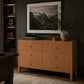 A warm tan leather six-drawer dresser in a styled home office setting, featuring brass knobs, a framed black and white mountain landscape print above it, and a potted plant on top.
