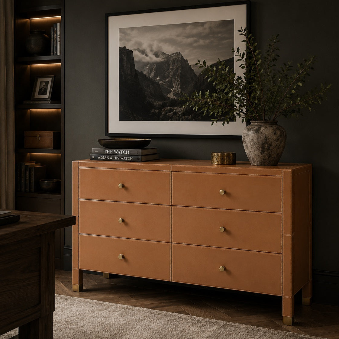 A warm tan leather six-drawer dresser in a styled home office setting, featuring brass knobs, a framed black and white mountain landscape print above it, and a potted plant on top.