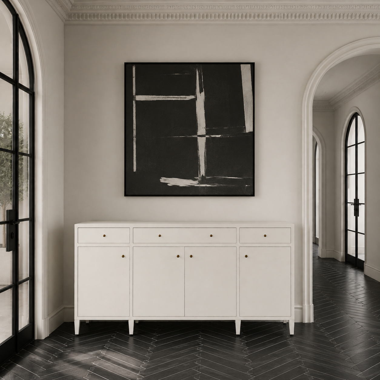 A white wooden sideboard cabinet with six drawers and three doors, accented by small brass knobs, positioned in a hallway below a black and white abstract painting. The room features a light gray wall, black herringbone wood floors, and arched windows on both sides.