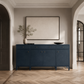 A navy blue wooden console cabinet with small brass knobs, styled in a modern entryway with a framed blank canvas on the wall, a vase with branches, and a black bowl on top, set against a beige wall and light wood herringbone floor.