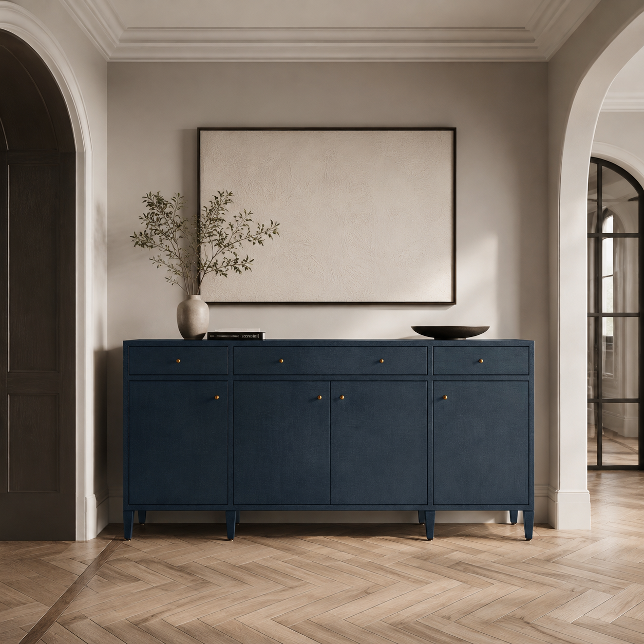 A navy blue wooden console cabinet with small brass knobs, styled in a modern entryway with a framed blank canvas on the wall, a vase with branches, and a black bowl on top, set against a beige wall and light wood herringbone floor.