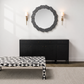 A modern interior scene featuring a black sideboard cabinet with brass hardware, a round gray wood-framed mirror on the wall, two gold wall sconces, and a black-and-white geometric patterned coffee table in the foreground.