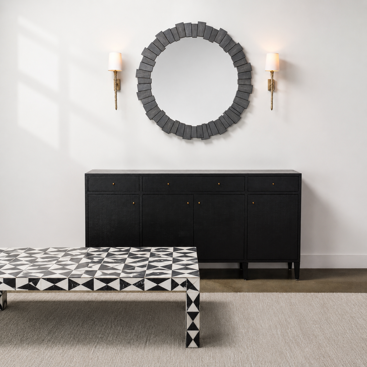 A modern interior scene featuring a black sideboard cabinet with brass hardware, a round gray wood-framed mirror on the wall, two gold wall sconces, and a black-and-white geometric patterned coffee table in the foreground.