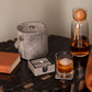 A styled tabletop scene showing a gray felted wool accessory set: a cylindrical covered container, a small square box with a ring holder, a glass tumbler with amber liquid and an ice cube, and a clear glass decanter with amber liquid and a wooden stopper. A book is partially visible on the left, and the products are arranged on a dark wood table against a warm beige wall.