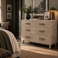 A beige three-drawer dresser in a styled bedroom setting, featuring dark metal bar handles, styled with a plant, books, and framed art on top.