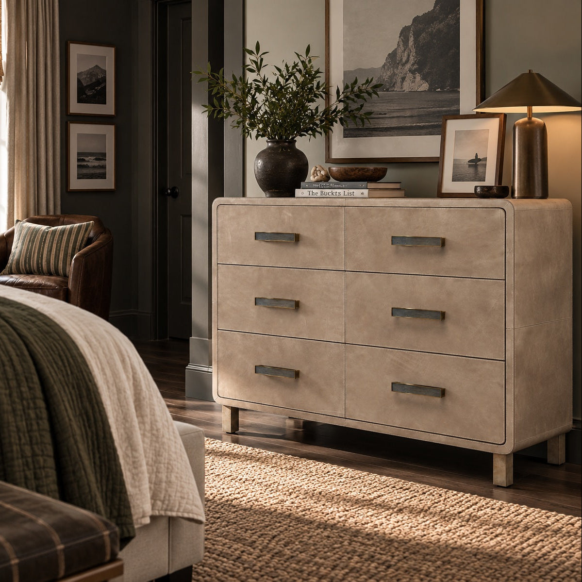 A beige three-drawer dresser in a styled bedroom setting, featuring dark metal bar handles, styled with a plant, books, and framed art on top.