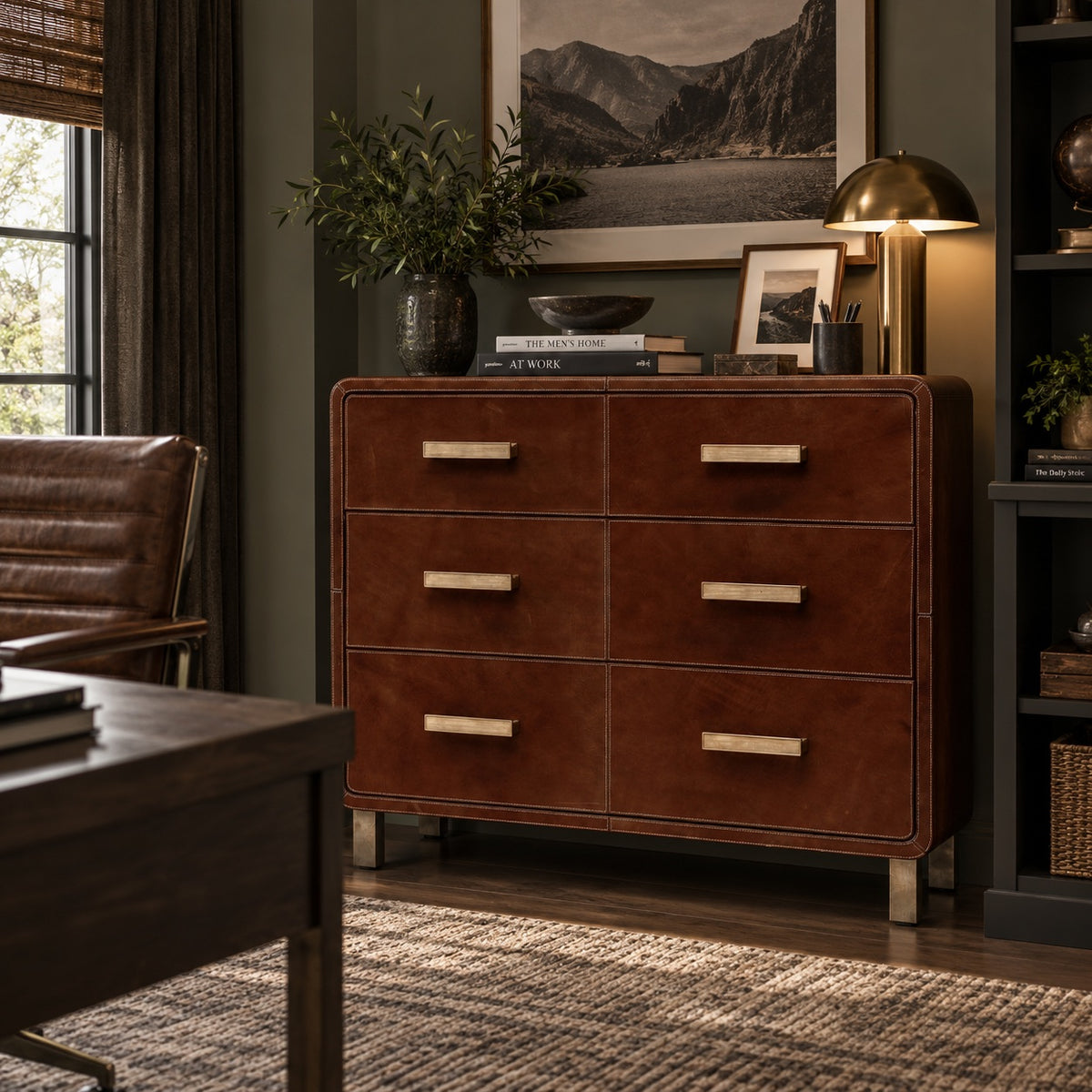 A mid-century modern chest of six drawers in warm reddish-brown leather or faux leather, with brass-toned rectangular handles, styled in a dark, elegant room next to a leather chair and under a framed landscape print.