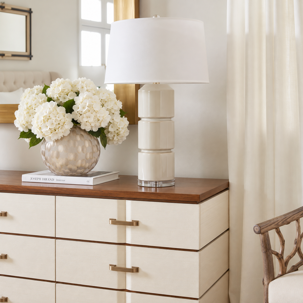 A cream-colored ceramic table lamp with a tiered stacked base and a white drum shade sits on a wooden dresser next to a vase of white flowers in a bright, modern room.