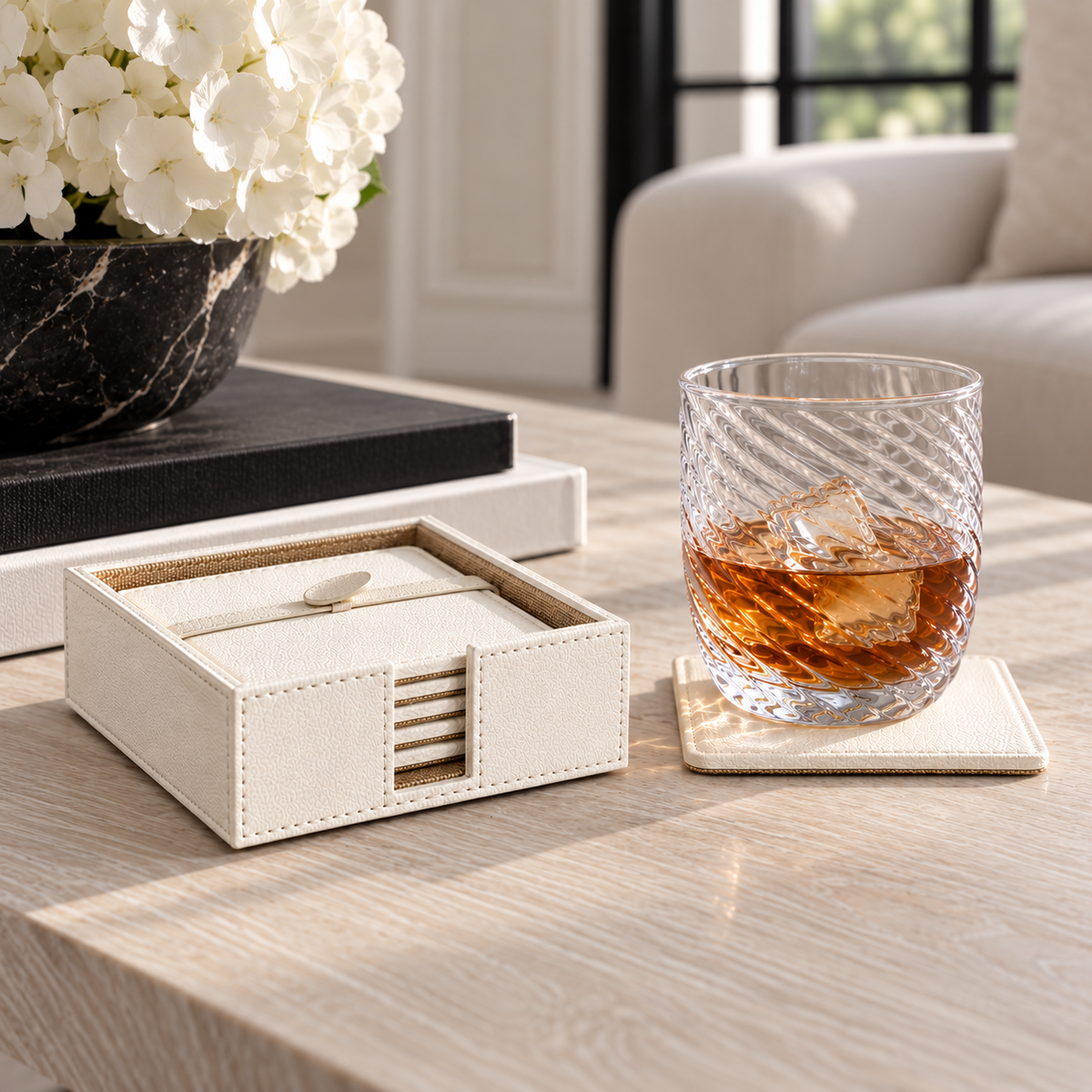 A cream-colored leatherette drink coaster set with a matching box and small spoon, styled on a light wood table next to a glass of whiskey on a coaster in a bright living room.