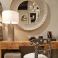 Elegant modern interior featuring a large round textured taupe wall mirror with a slim gold frame above a burl wood desk with brass knobs. A sculptural fluted stone table lamp with an ivory shade complements a black and ivory floral inlay chair, creating a sophisticated, neutral luxury space.