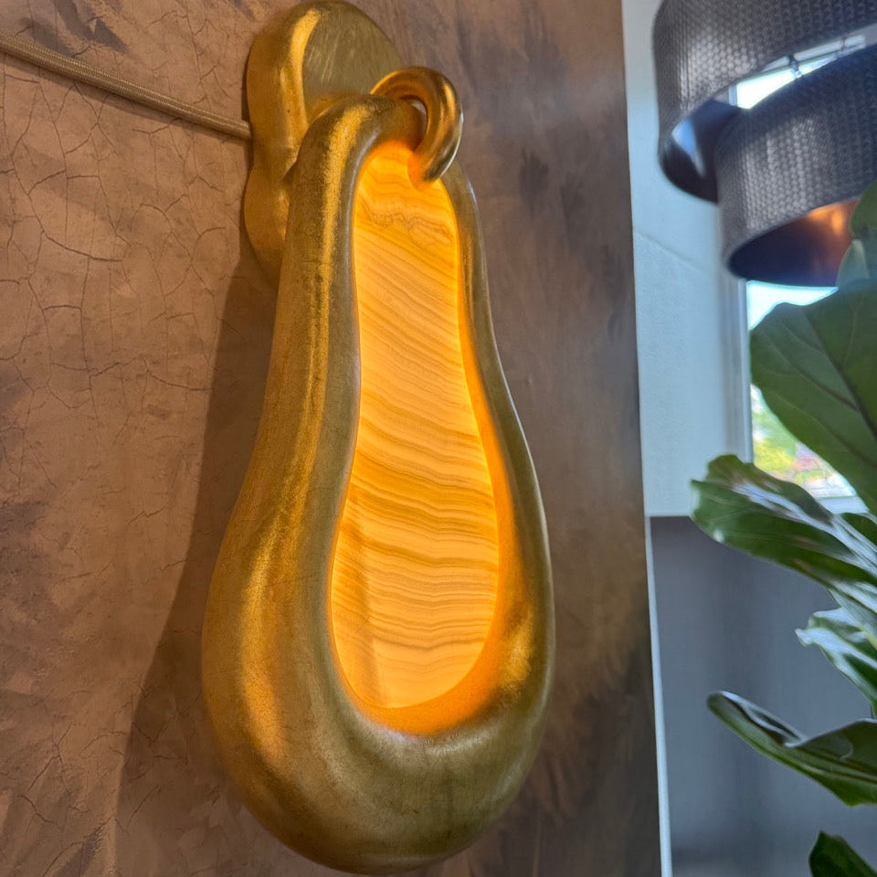 A sculptural gold wall sconce with a teardrop-shaped metallic body and a glowing warm yellow-orange interior, mounted on a brown textured wall with part of a window and green leaves visible at the side.