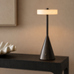 A modern table lamp with a dark bronze cone-shaped base and a glowing cylindrical shade, placed on a dark wood console table against a warm beige wall.