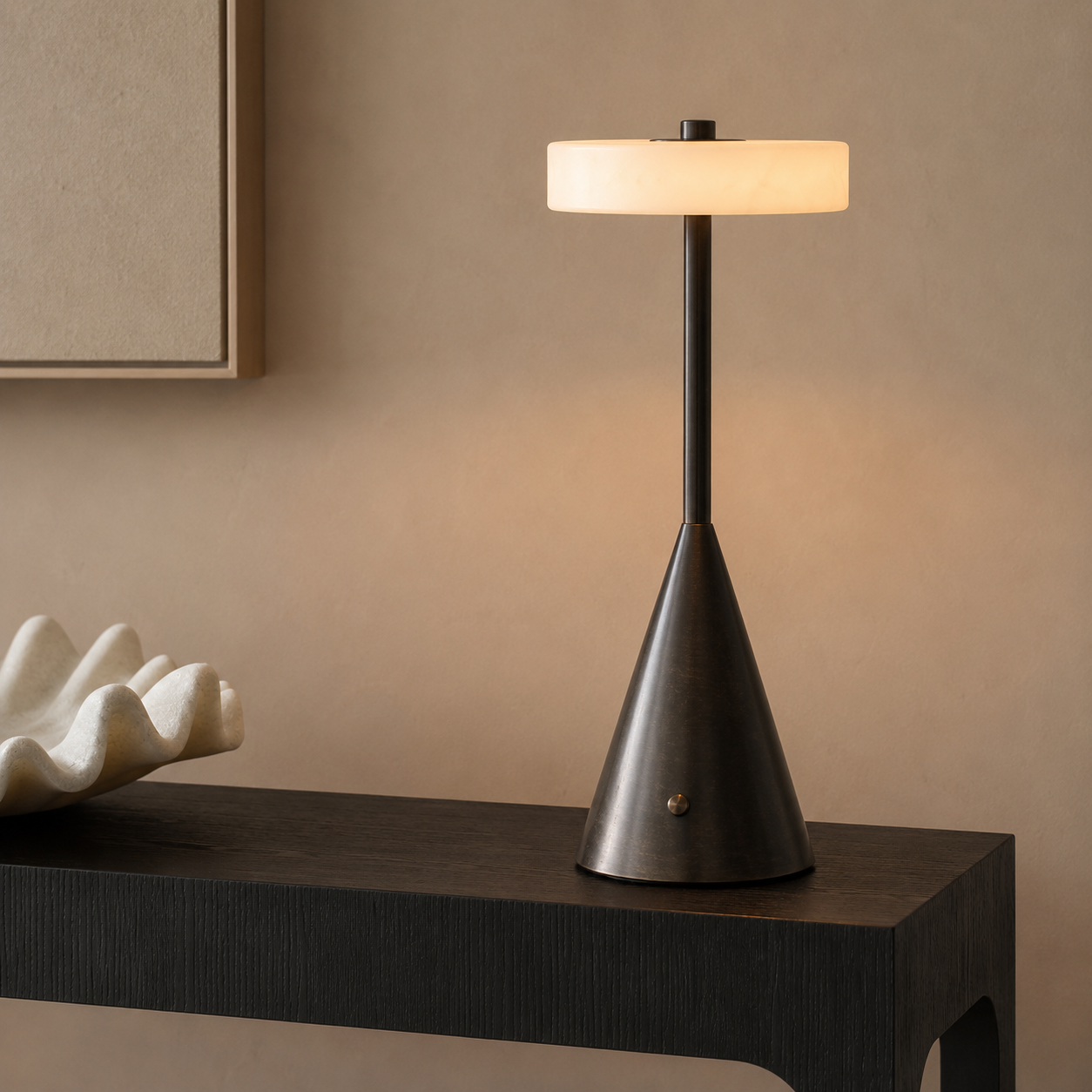A modern table lamp with a dark bronze cone-shaped base and a glowing cylindrical shade, placed on a dark wood console table against a warm beige wall.