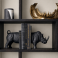 Black rhinoceros bookends holding a row of books on a dark wooden shelf, with a gold shark fin sculpture on the top shelf and a black and white canister on the left.