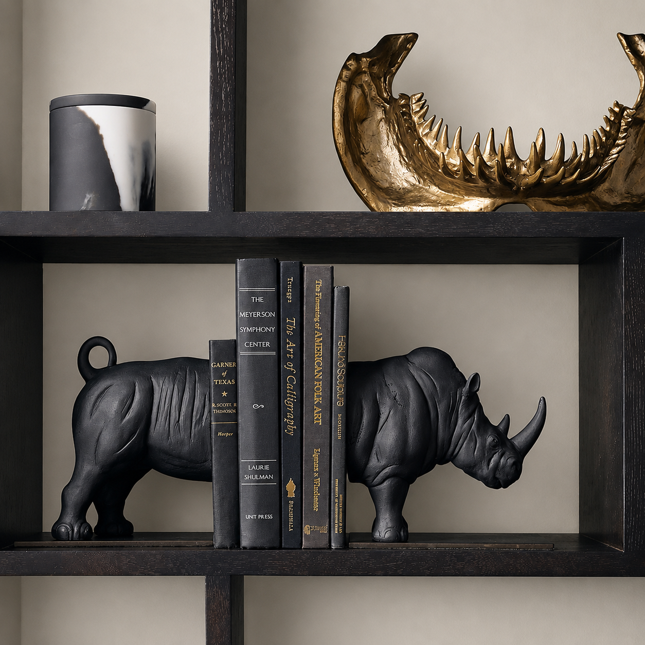 Black rhinoceros bookends holding a row of books on a dark wooden shelf, with a gold shark fin sculpture on the top shelf and a black and white canister on the left.