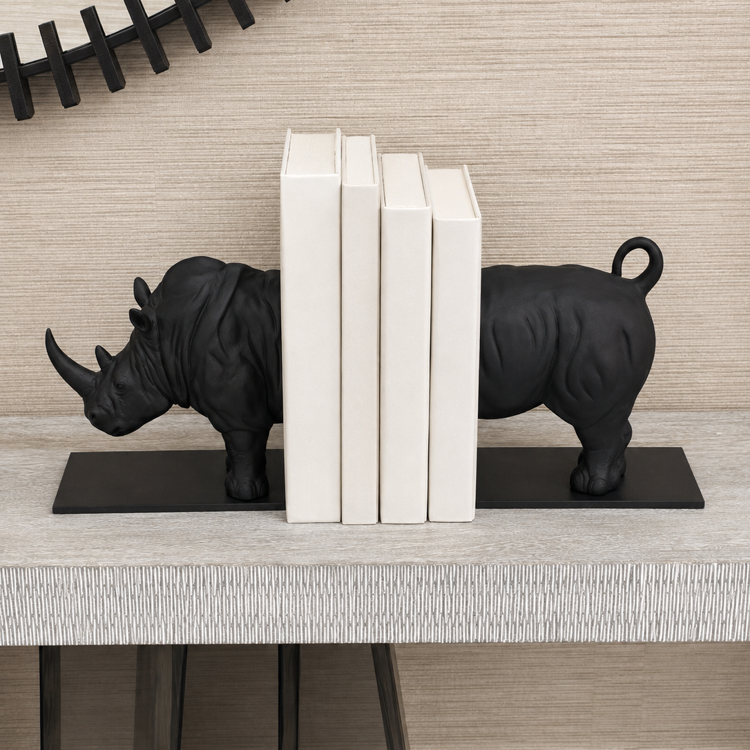 Black rhinoceros bookends holding up four white books on a light gray textured table against a beige wood-paneled wall.