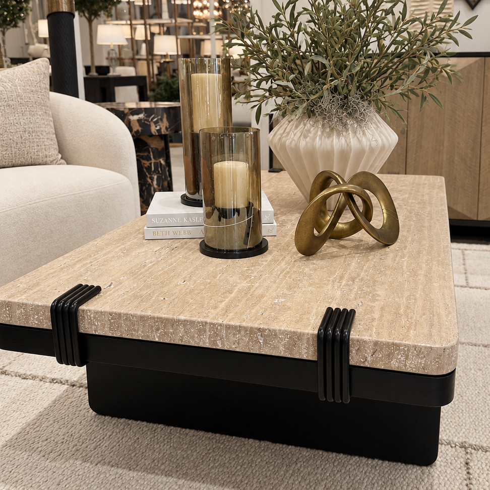 A modern square coffee table with a beige textured stone top and a black metal base, styled with candles, a vase of greenery, and a decorative sculpture in a showroom setting.