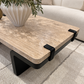 A modern rectangular coffee table with a beige travertine stone top and a black metal frame, shown in a living room setting with a sofa, rug, and a white vase on top.