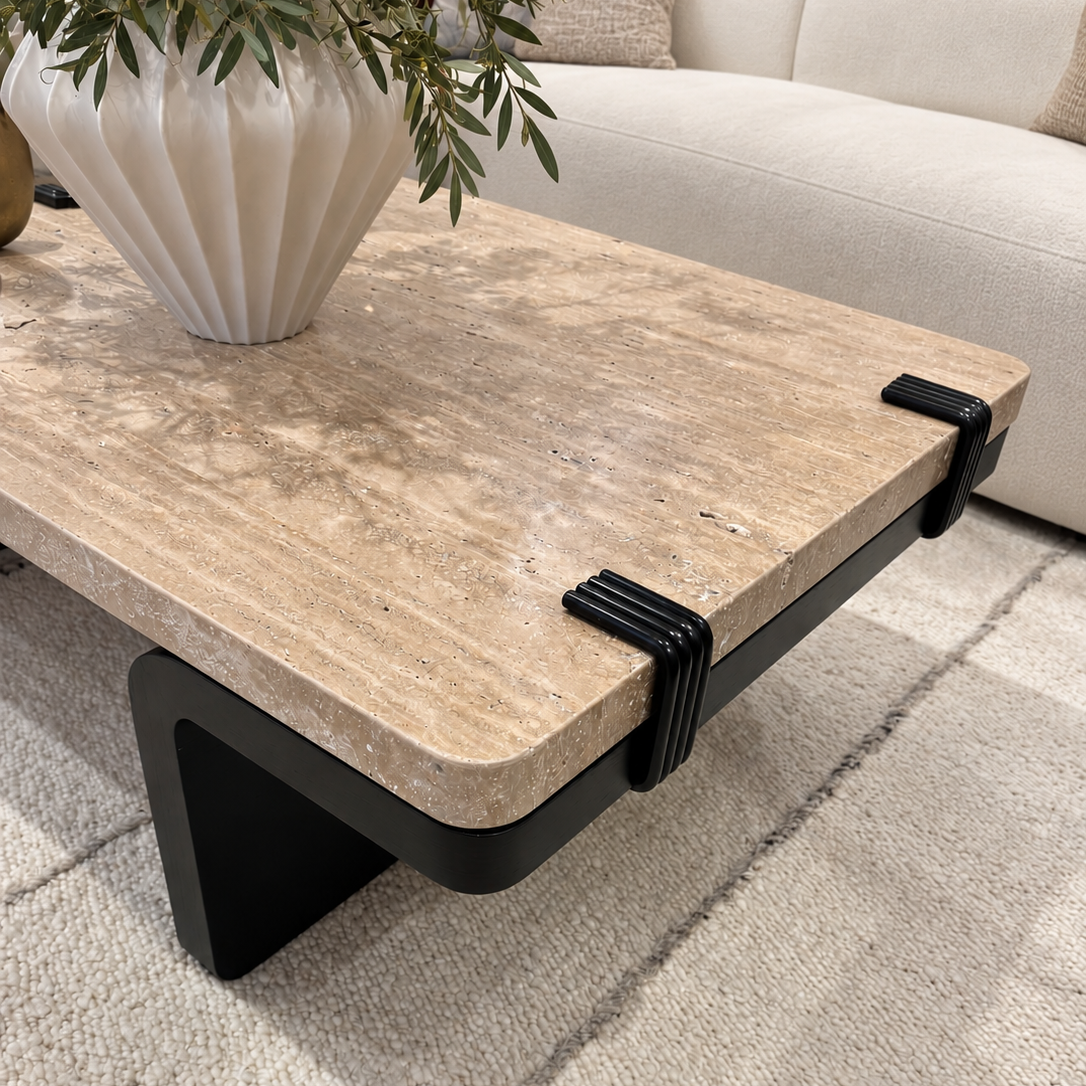 A modern rectangular coffee table with a beige travertine stone top and a black metal frame, shown in a living room setting with a sofa, rug, and a white vase on top.