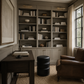 A styled home office corner featuring a large built-in bookcase in a muted gray-green tone, filled with books and decor. A dark wood desk sits on the left, a black cylindrical ottoman in the foreground, and a brown leather armchair is positioned by a window with beige curtains.