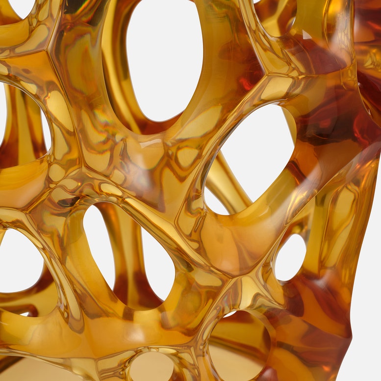 Close-up of a translucent amber-gold sculptural object with an organic, web-like open structure against a white background.