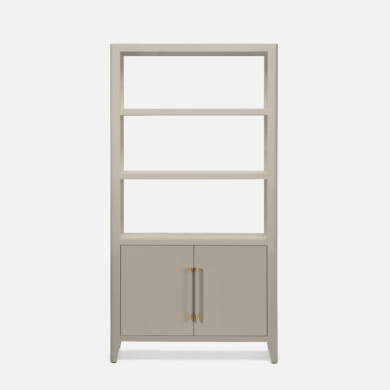 A tall, narrow bookcase with four open shelves on the upper portion and two cabinet doors on the lower portion, finished in a light warm gray or greige tone with slim gold-toned vertical handles, shown on a plain light gray background.