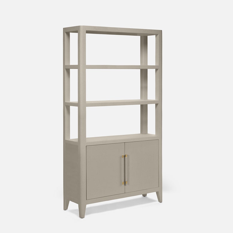 A tall, narrow bookcase with four open shelves on the upper portion and two cabinet doors on the lower portion, finished in a light warm gray or greige tone with slim gold-toned vertical handles, shown on a plain light gray background.