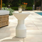 A white ribbed pedestal side table with a wide flared top, shown outdoors on a stone patio with a glass of water on top.