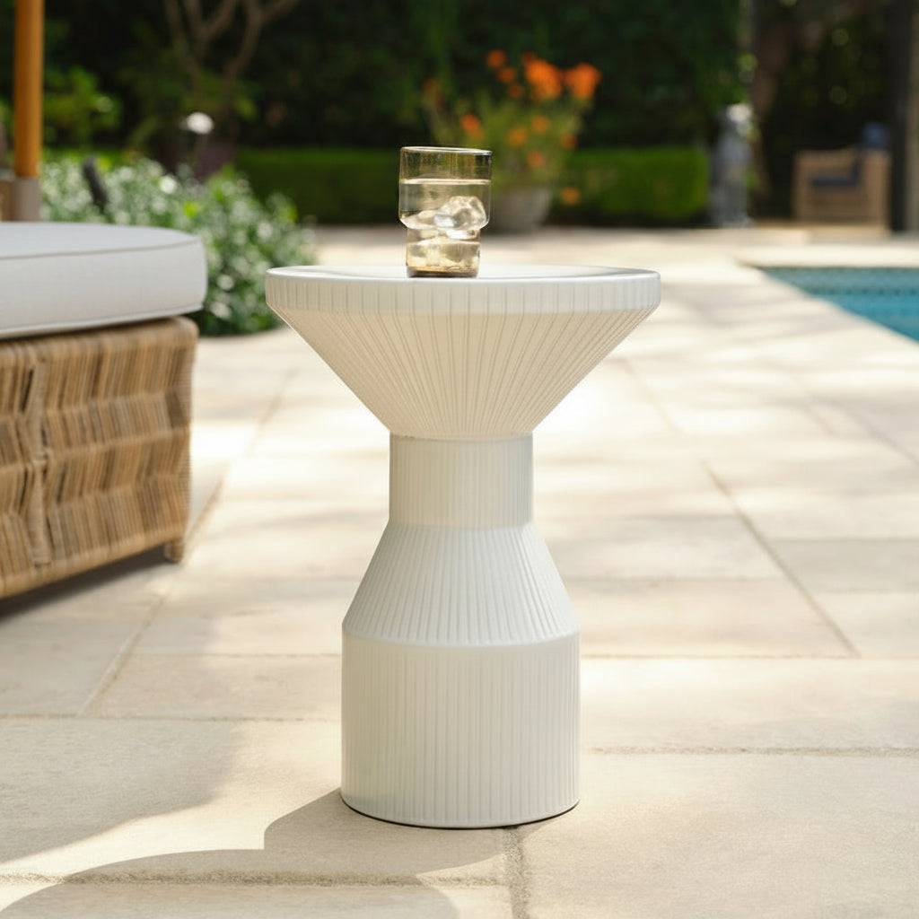 A white ribbed pedestal side table with a wide flared top, shown outdoors on a stone patio with a glass of water on top.