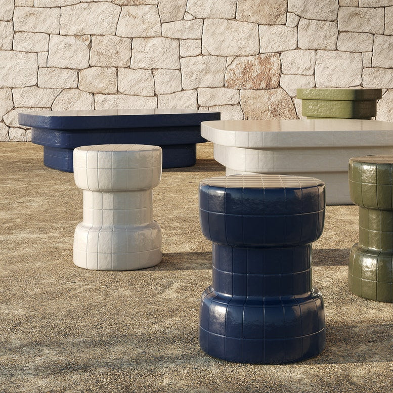 A set of modern stacked barrel stools in glossy navy blue, matte white, and olive green, arranged on a textured stone floor in front of a rustic stone wall.