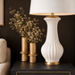 A white ceramic table lamp with a fluted ribbed body and gold metal accents sits on a dark side table next to two gold vases with dried white flowers against a dark wood wall.