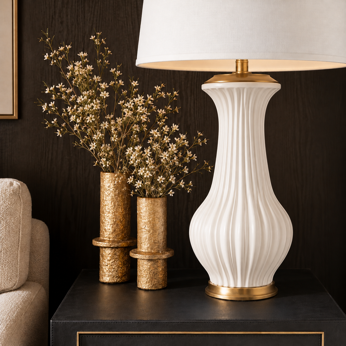 A white ceramic table lamp with a fluted ribbed body and gold metal accents sits on a dark side table next to two gold vases with dried white flowers against a dark wood wall.