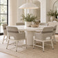 A round white dining table surrounded by six upholstered armchairs in a bright, modern dining room. The chairs feature textured light beige and taupe woven backs with white seat cushions.