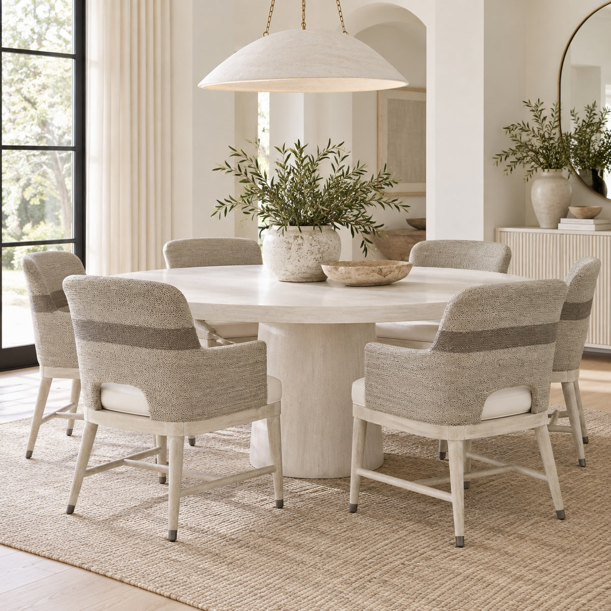 A round white dining table surrounded by six upholstered armchairs in a bright, modern dining room. The chairs feature textured light beige and taupe woven backs with white seat cushions.