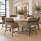 A dining room scene with a round white dining table and six woven armchairs with gray metal frames and dark blue horizontal stripe details on the backrests. The chairs are arranged around the table on a natural jute rug, with a large white dome pendant light hanging above.