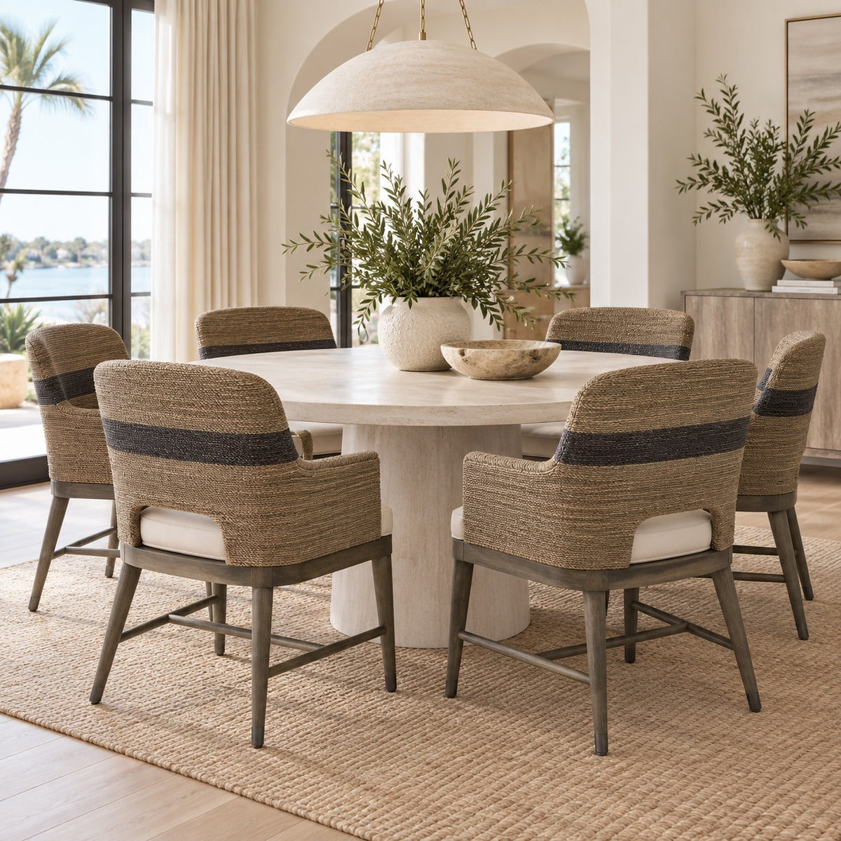 A dining room scene with a round white dining table and six woven armchairs with gray metal frames and dark blue horizontal stripe details on the backrests. The chairs are arranged around the table on a natural jute rug, with a large white dome pendant light hanging above.